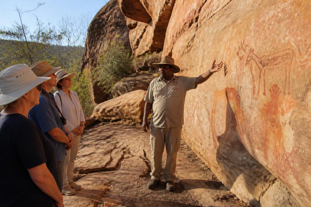 An Indigenous guide standing in front of ancient rock art formations on weathered sandstone, gesturing toward ochre-coloured hand stencils and animal figures while a small group of mature travellers listen intently, soft morning light casting long shadows across the gallery wall