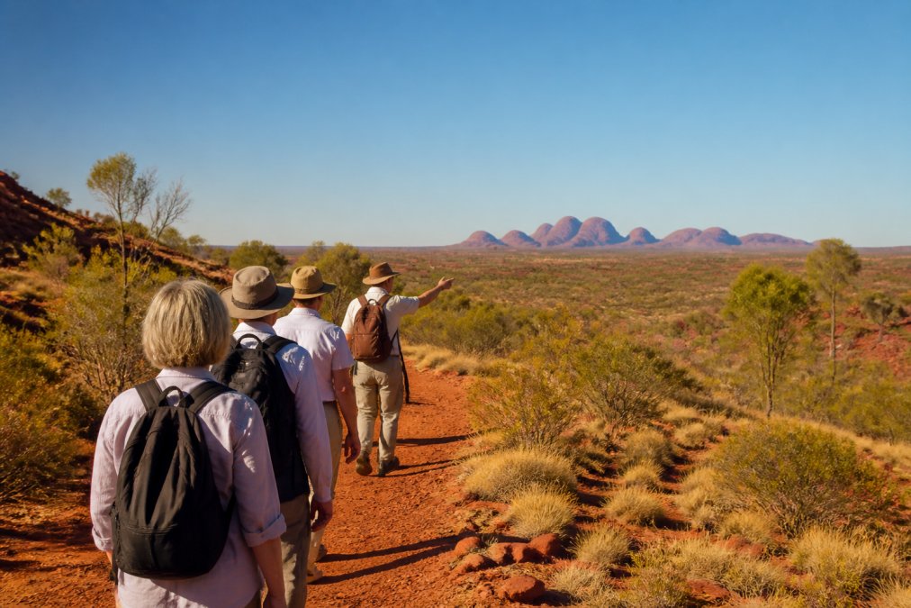 A small group of mature travellers walking single-file along a red-dirt trail through sparse desert scrubland, a guide in front pointing toward distant rock formations, clear blue sky overhead and golden light indicating late afternoon, the landscape stretching into the distance