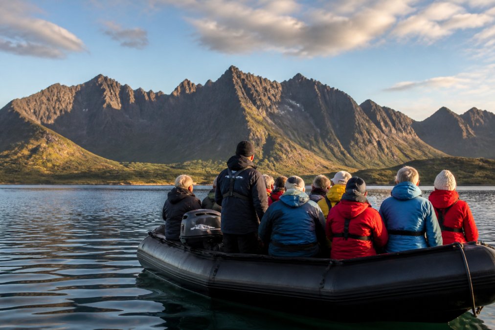 Group of visitors in a boat on calm waters with dramatic rocky Cuillin peaks rising in the distance, golden afternoon light illuminating the mountainous landscape