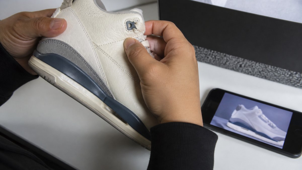 Hands comparing sneaker stitching and box labels against a digital reference photo to verify authenticity
