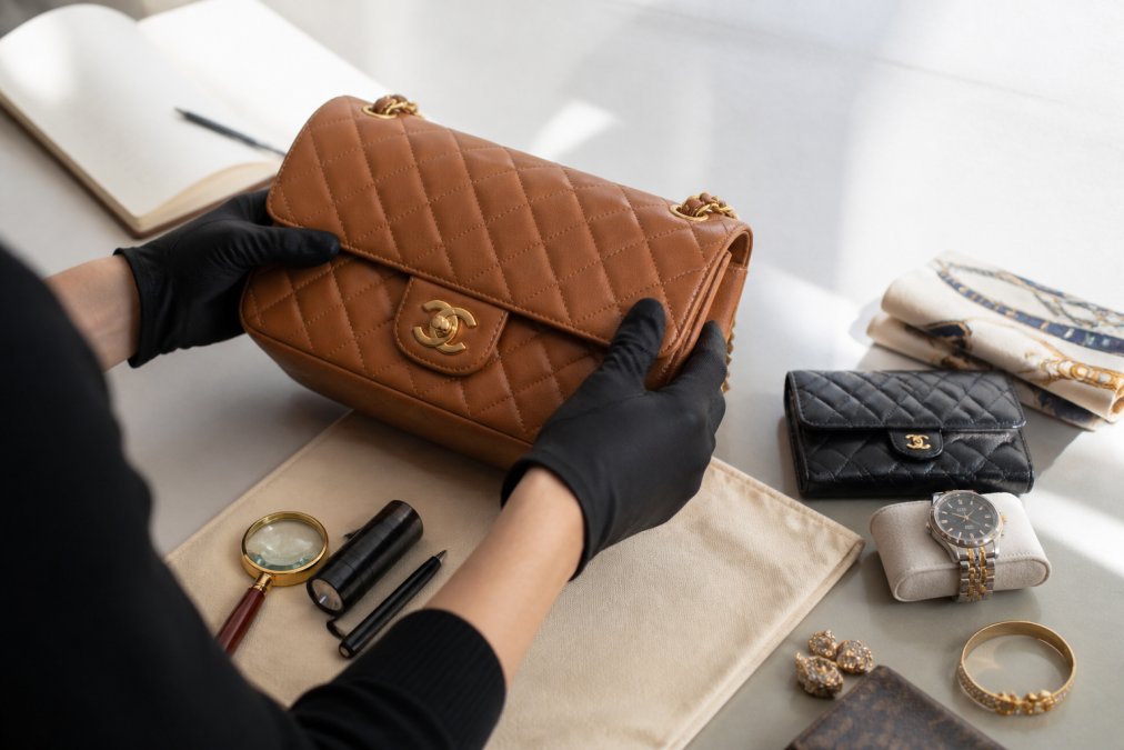Hands carefully examining pre-owned luxury handbag leather for patina, wear patterns, and condition with natural side lighting on neutral background
