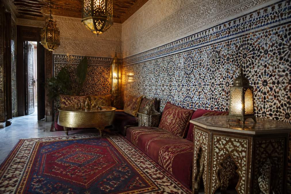 Richly decorated Moroccan interior featuring intricate zellige tilework in geometric patterns covering walls, hand-woven area rug with traditional motifs in deep jewel tones, ornate brass lanterns hanging from carved wooden ceiling, low seating with layered textile cushions in burgundy and gold, and carved wooden side table with mother-of-pearl inlays catching warm lamplight