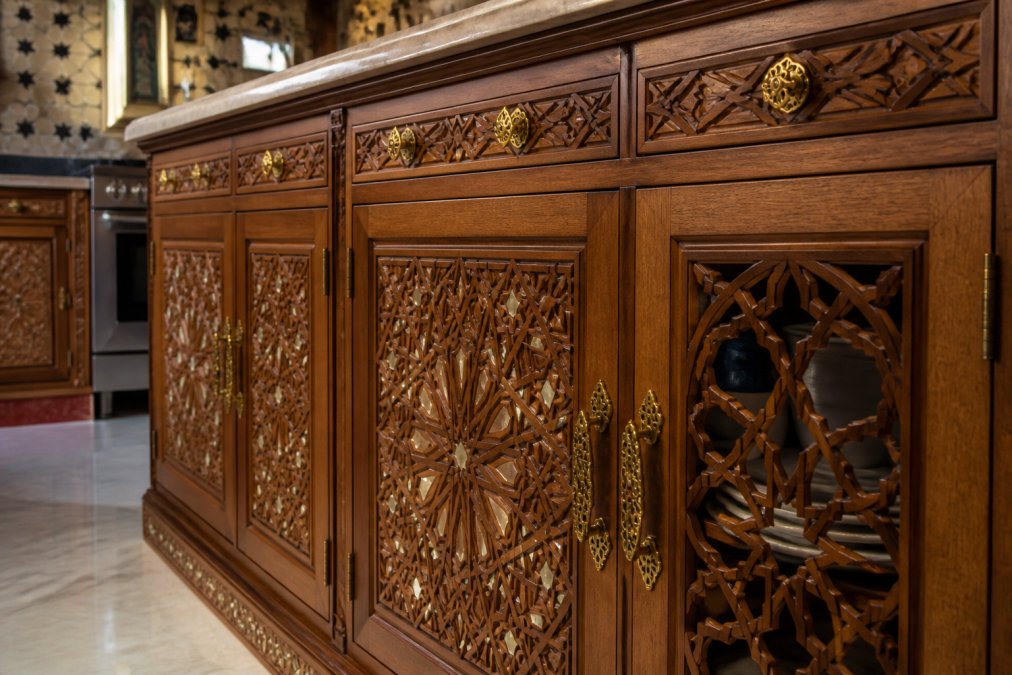 Close-up of hand-carved Moroccan kitchen cabinetry featuring intricate geometric star and diamond patterns in cedar wood, mother-of-pearl inlays catching warm light, hand-forged brass hardware with traditional patterns, and carved wooden grille detail revealing interior shelving