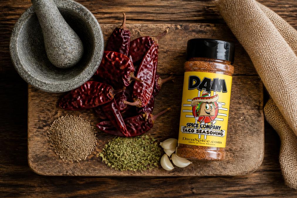 Overhead view of dried red New Mexico chiles, whole cumin seeds, Mexican oregano leaves, and garlic cloves arranged on a weathered wooden cutting board with a traditional molcajete and pestle