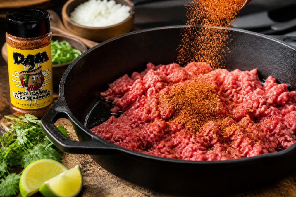 Close-up of vibrant red seasoning being sprinkled over raw ground beef in a cast iron skillet with oil shimmering, fresh lime wedges and cilantro sprigs visible at the edge of the pan