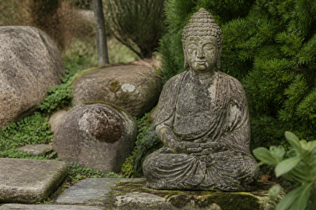Frost-proof stone Buddha statue positioned in serene outdoor garden setting surrounded by natural stone elements, with visible weathering and aged patina from environmental exposure