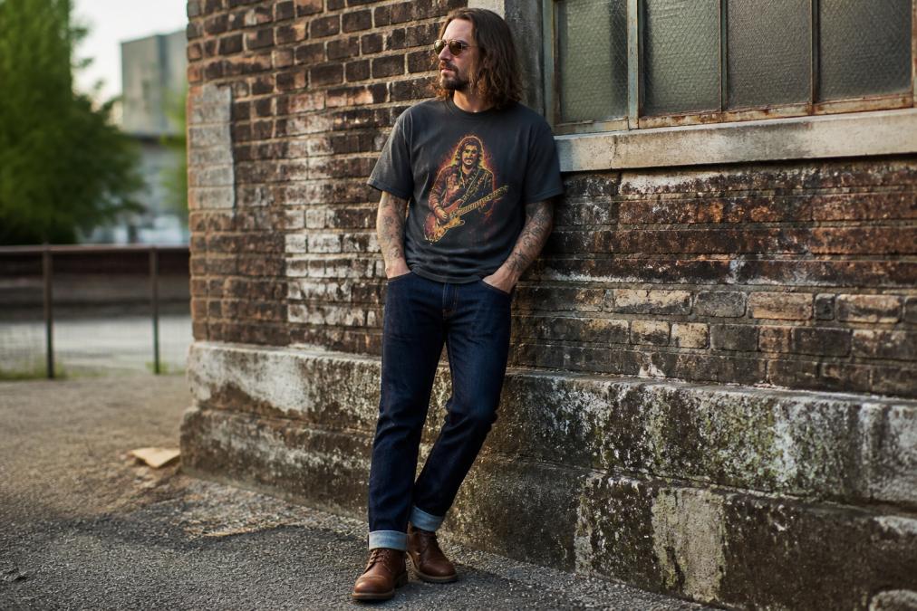 Person wearing a heavyweight garment-dyed graphic tee featuring classic rock imagery, styled with dark raw denim jeans and worn leather boots, demonstrating the boxy fit and soft hand-feel of an authentic vintage look in natural daylight