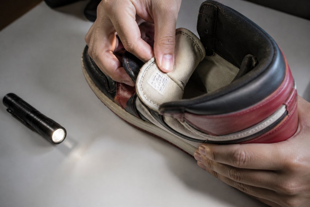 Close-up of hands inspecting the inner size tag and stitching on a pre-owned high-top sneaker on a clean table, with a phone flashlight nearby