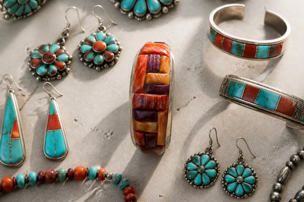 Curated display of Zuni earrings and bracelets arranged on a neutral surface, showcasing petite point and inlay techniques with various stone colors including turquoise, coral, and jet, with natural lighting