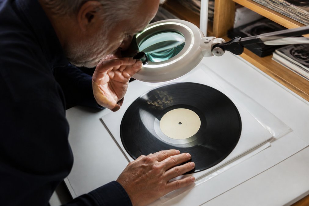 Professional examiner inspecting signed vinyl record under specialized magnification lamp, studying autograph details with forensic precision on white examination table
