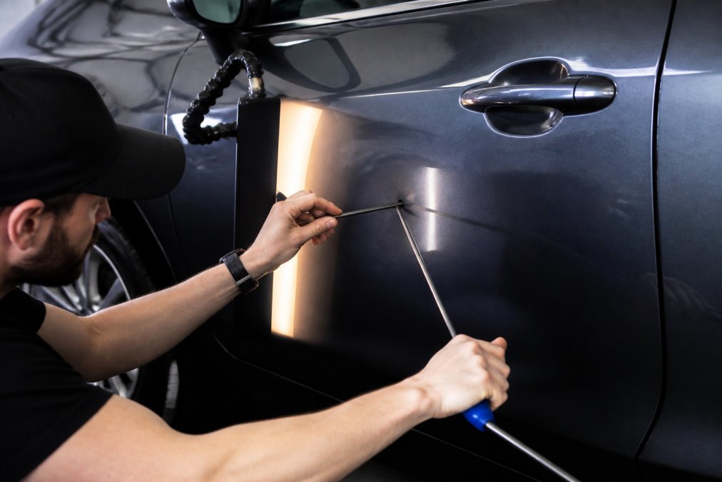 Professional technician using specialized rods and hooks on vehicle door panel to remove a door ding, showing precision technique with metal memory working, hands-on demonstration in well-lit workshop