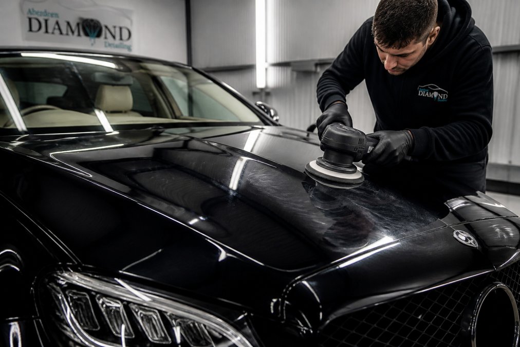 Close-up of a detailer using a dual-action polishing machine on the bonnet of a dark-coloured prestige car inside a professional workshop, with studio lighting revealing the paintwork clarity
