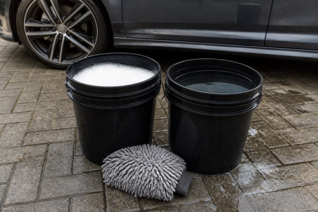 Close-up van twee emmers water naast elkaar met een microvezelwashandschoen, één emmer met zeepachtig water en één met schoon water, op een oprit bij een auto