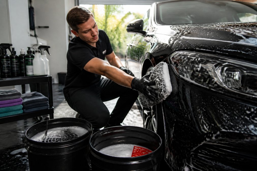 Persoon die een auto wast met twee emmers, microvezeldoeken en professionele reinigingsproducten in een thuisgarage met natuurlijk daglicht