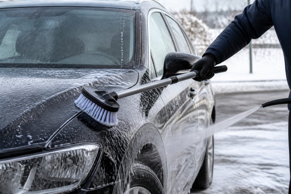 Iemand die een natte auto met een zachte borstel en zeepschuim wast in een winteromgeving, met zichtbare waterstraal en zichtbare waterdruppels op het voertuig