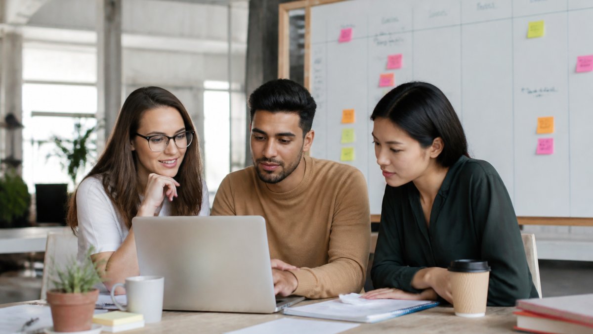 A small startup team of three people reviewing an onboarding checklist on a laptop in a bright open-plan office, with a whiteboard showing a hiring timeline behind them