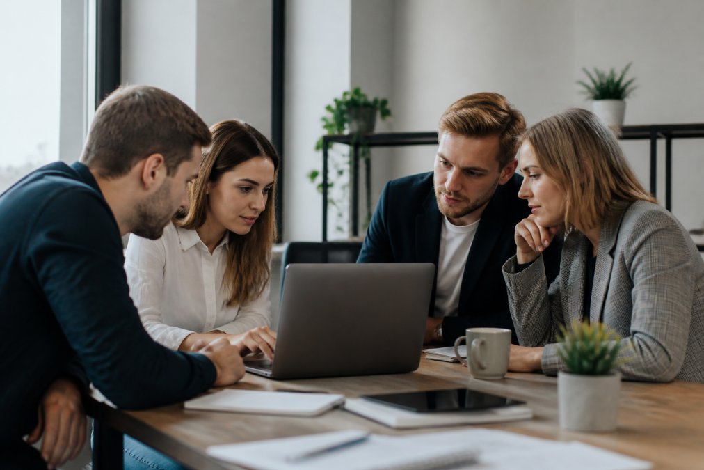 A marketing team collaborating around a laptop, reviewing keyword data and workflow automation diagrams on screen in a modern office setting