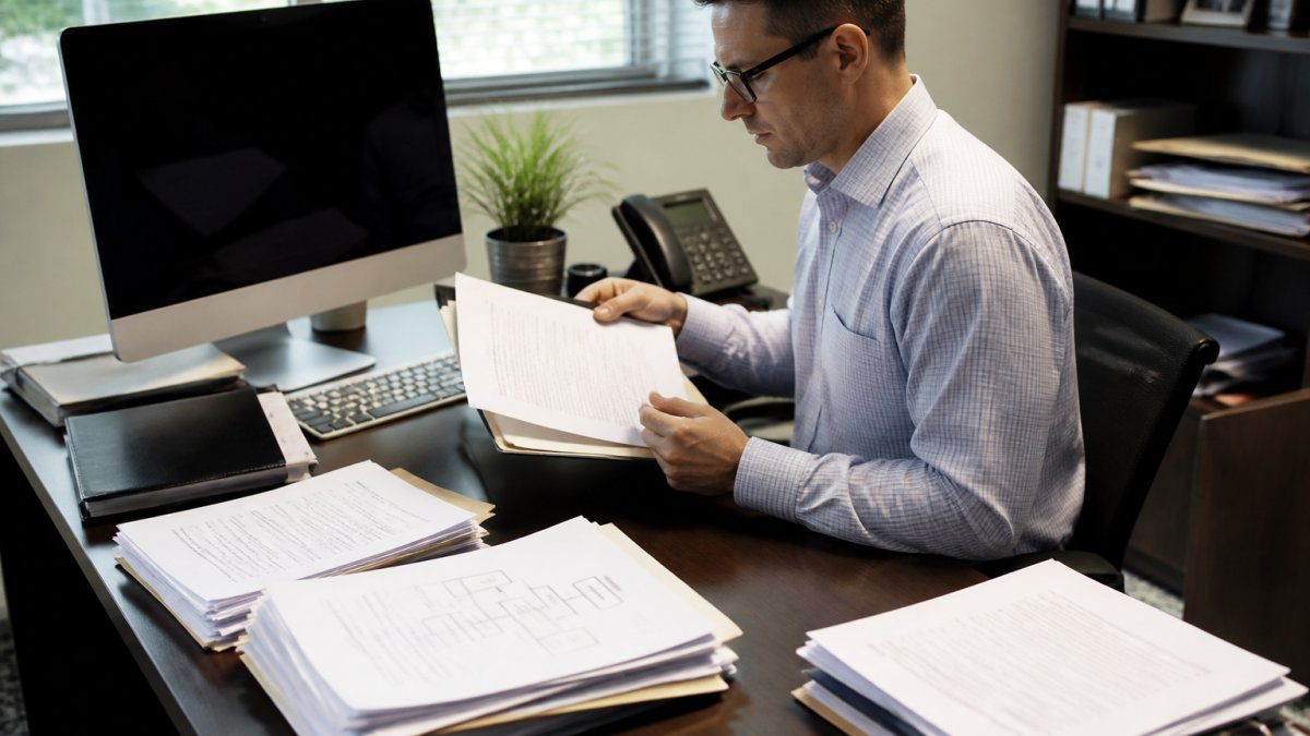 Professional accountant reviewing and organizing multiple legal entity documents and ownership structures at a desk with computer showing centralized entity records database