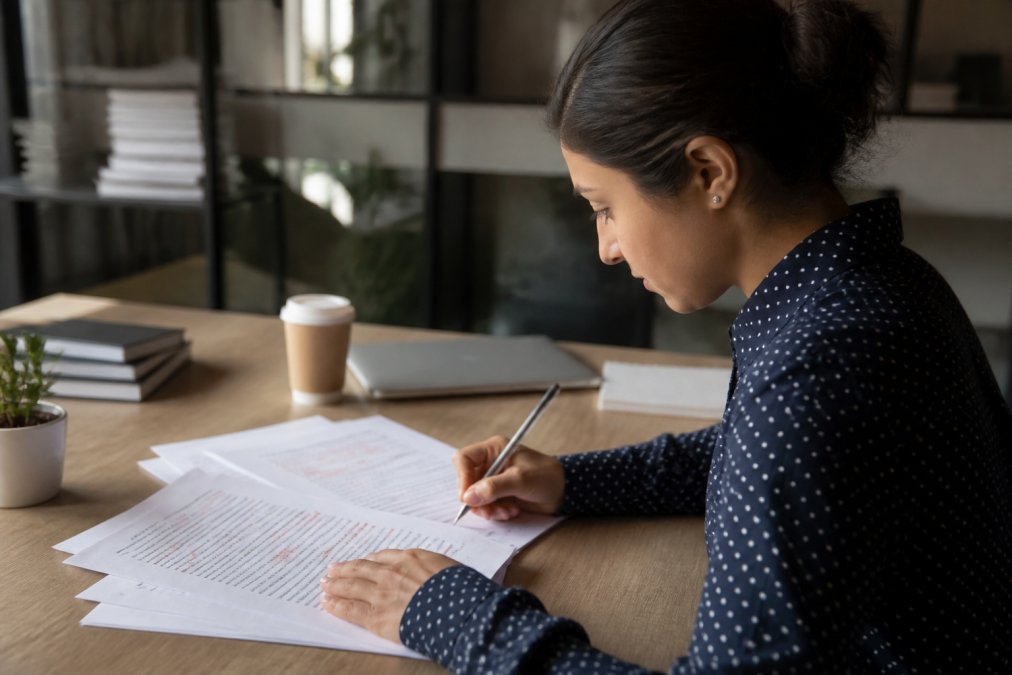 Marketer reviewing and editing AI-generated content on computer screen in modern office setting, with red editing marks visible on document