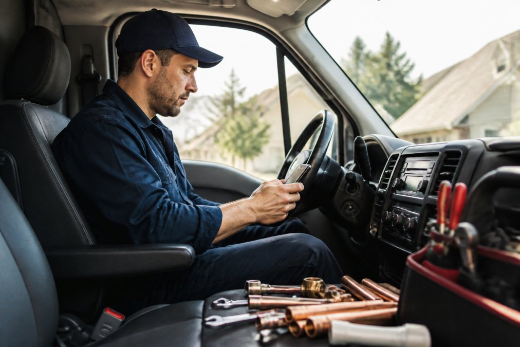 Plumbing technician in work vehicle checking phone for routed job assignments with real-time lead notifications visible on screen, natural daylight through windshield