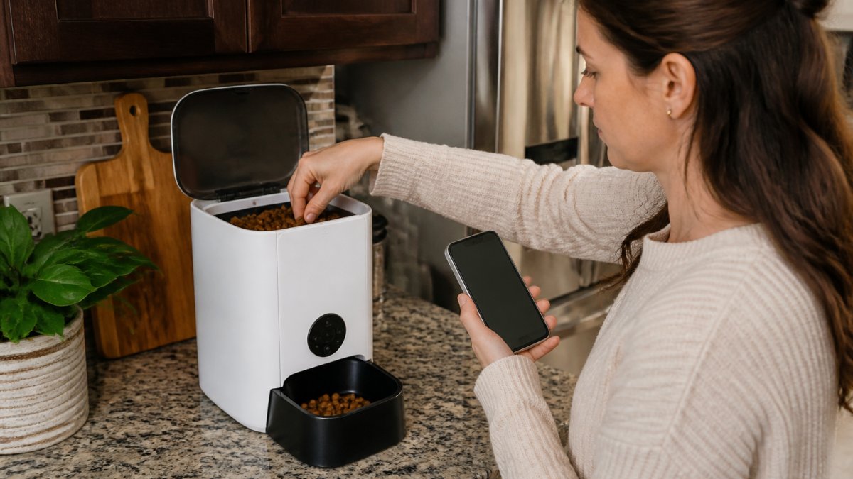 Pet owner checking an automatic feeder's food hopper and testing offline functionality at home before a trip, with the feeder sitting on a kitchen counter and the owner holding a smartphone showing the feeder app