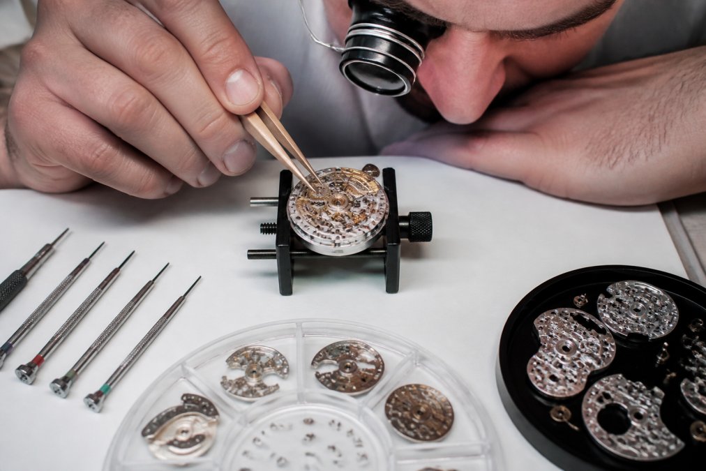 A watchmaker's hands using fine-tipped tweezers to position a tiny gear component on a white-lit workbench, with a partially assembled caliber, small screwdrivers, and movement parts arranged in organized trays nearby