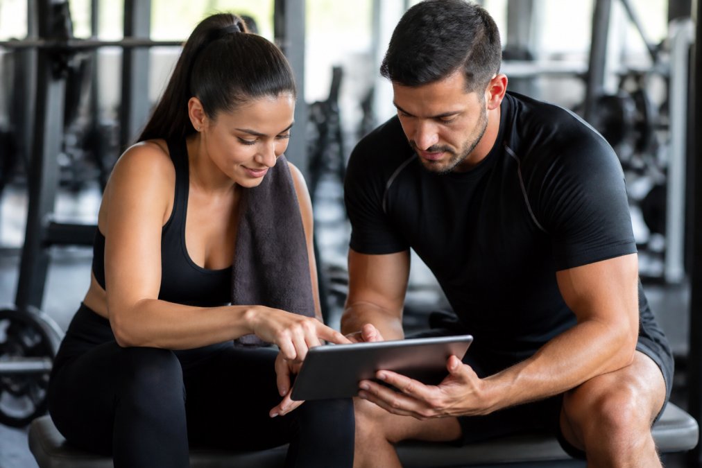 Coach sportif et client consultant ensemble un tableau de progression sur une tablette dans un environnement de coaching moderne, avec des graphiques de suivi automatisé visibles à l'écran