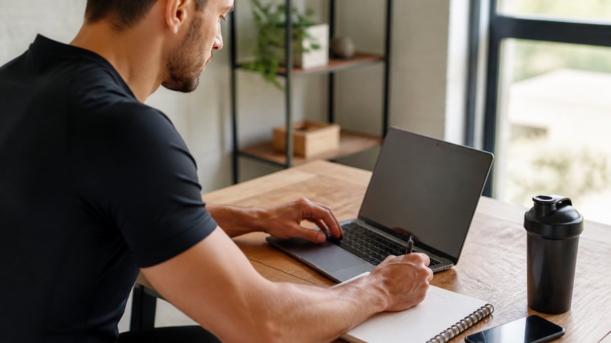 Un coach sportif assis à son bureau devant un ordinateur portable affichant une interface de planification d'entraînement, carnet de notes et téléphone posés à côté, dans un espace de travail lumineux