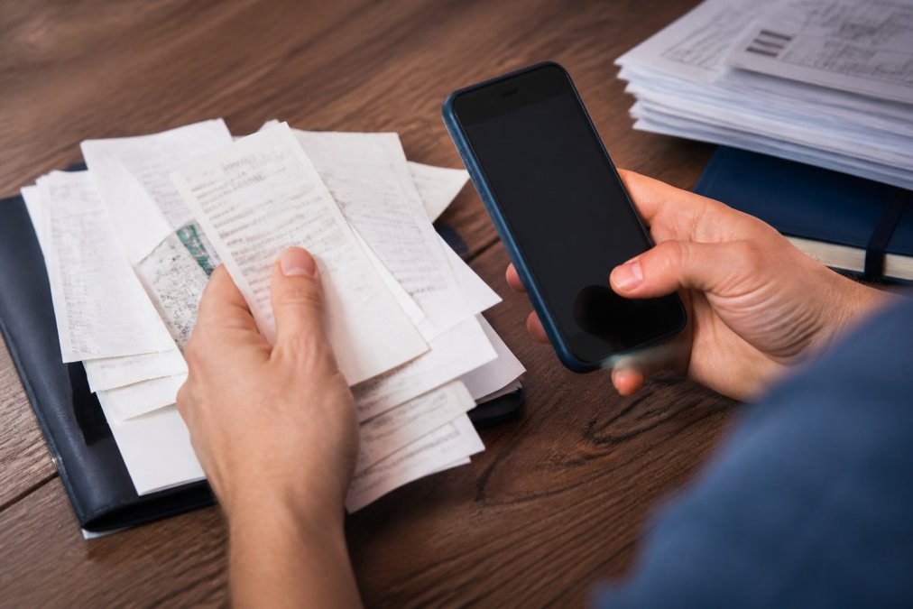 Close-up of hands organizing printed service receipts and documents next to a smartphone displaying a digital maintenance app with organized service history records