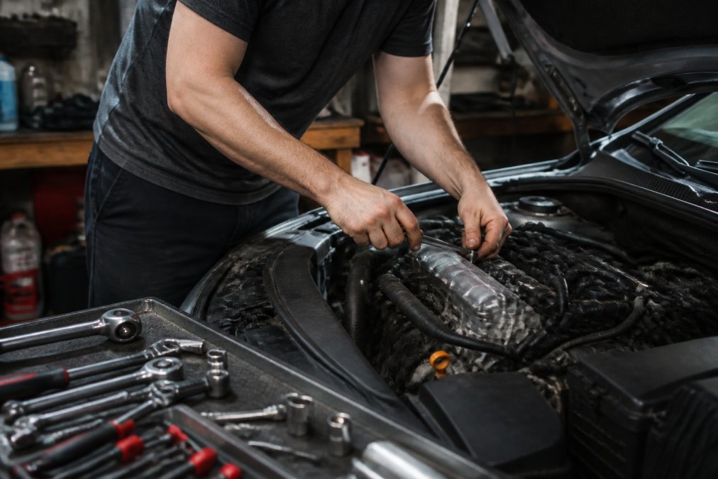 Hands performing DIY car maintenance on an engine component in a home garage setting, with tools scattered on the workbench and engine compartment visible