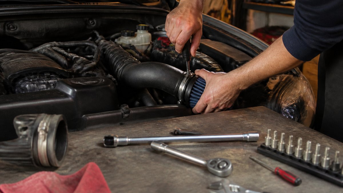 A mechanic's hands tightening a cold air intake clamp in a home garage, tools scattered on a workbench, car engine bay visible in the background under warm overhead lighting