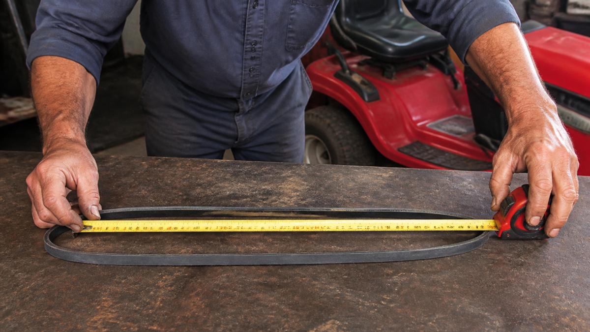 Close-up of a mechanic's hands using a tape measure on a riding mower's deck belt, showing proper measurement technique with the belt laid flat on a workbench under natural light