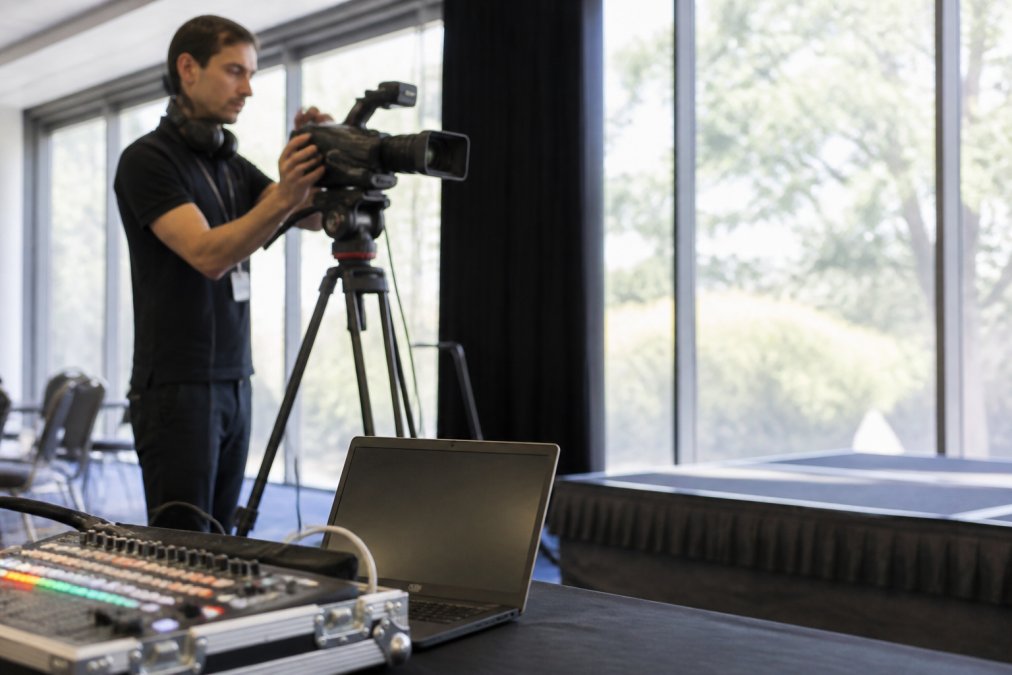 An AV technician adjusting a camera on a tripod in a conference room, with a mixing desk and laptop visible in the foreground, natural light from windows