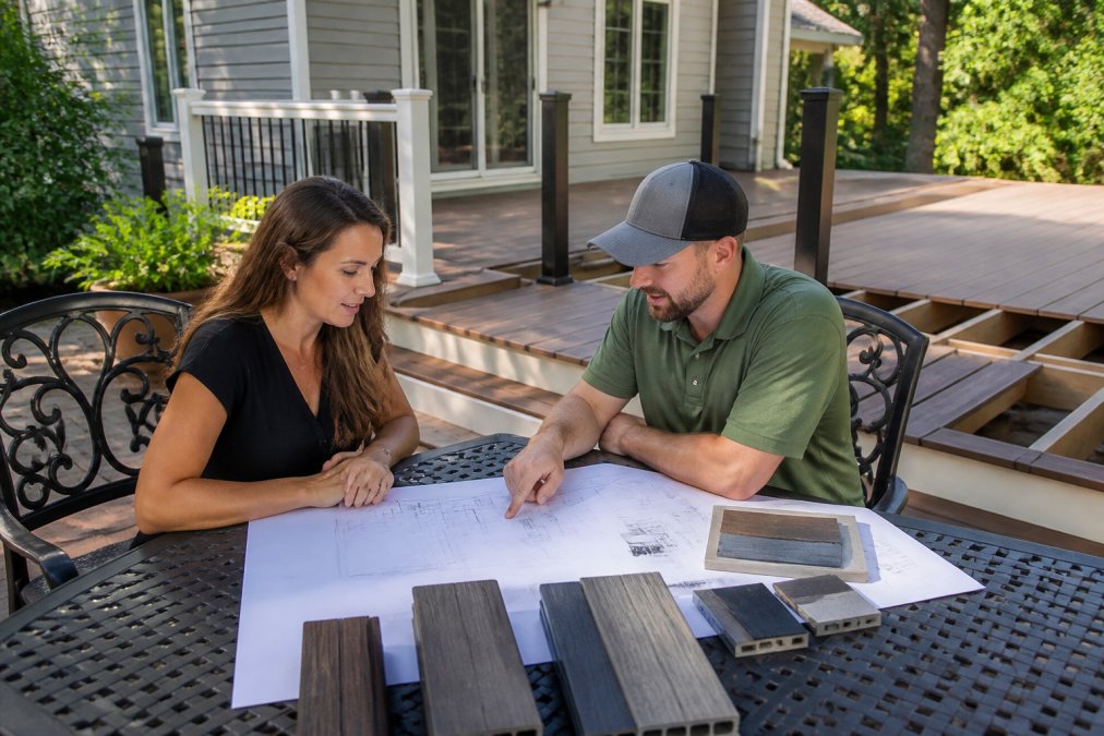 A homeowner and deck contractor reviewing custom deck blueprints and material samples on a backyard patio table, with a partially built composite deck visible in the background