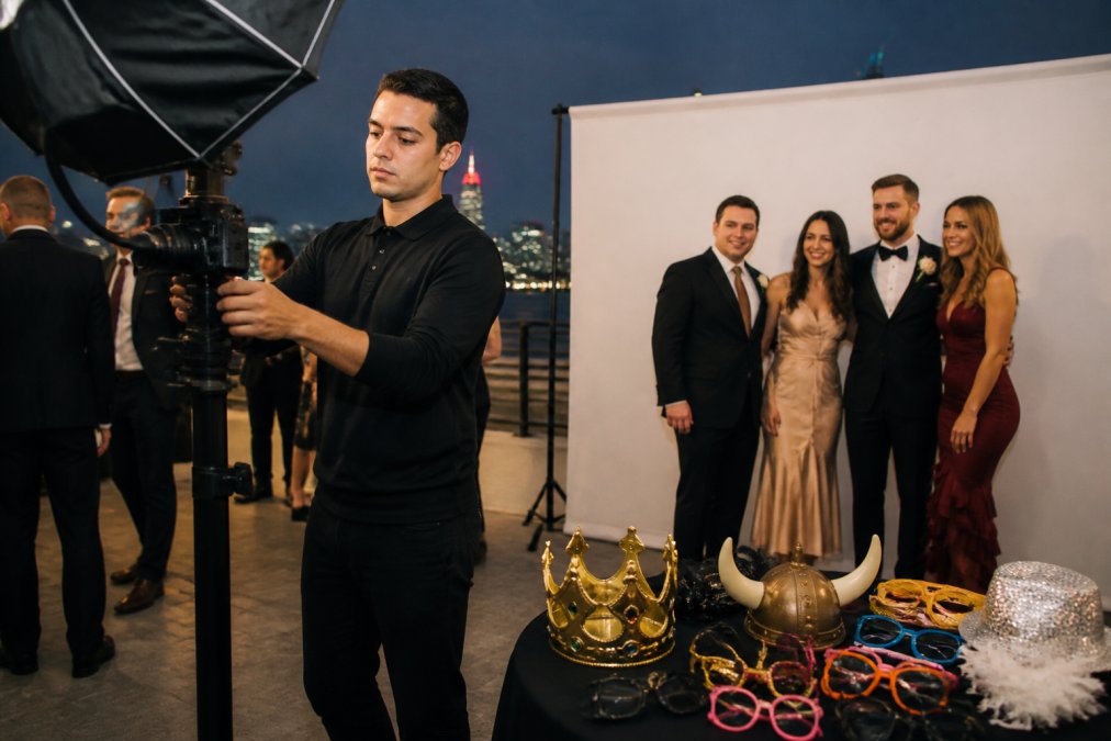 A photo booth attendant in a black shirt adjusting a professional camera and studio lighting inside an open-air booth at an evening event, with a custom branded backdrop and a small table of props visible in the foreground A photo booth attendant in a black shirt adjusting a professional camera and studio lighting inside an open-air booth at an evening event, with a custom branded backdrop and a small table of props visible in the foreground