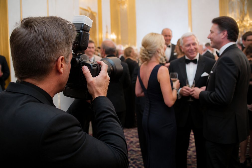 Professional photographer capturing candid moments at a corporate gala, with elegantly dressed attendees networking in the background, photographer using professional camera equipment