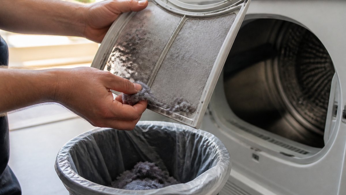 Close-up of hands cleaning a dryer lint screen over a trash can, showing accumulated lint fibers and proper removal technique in natural daylight
