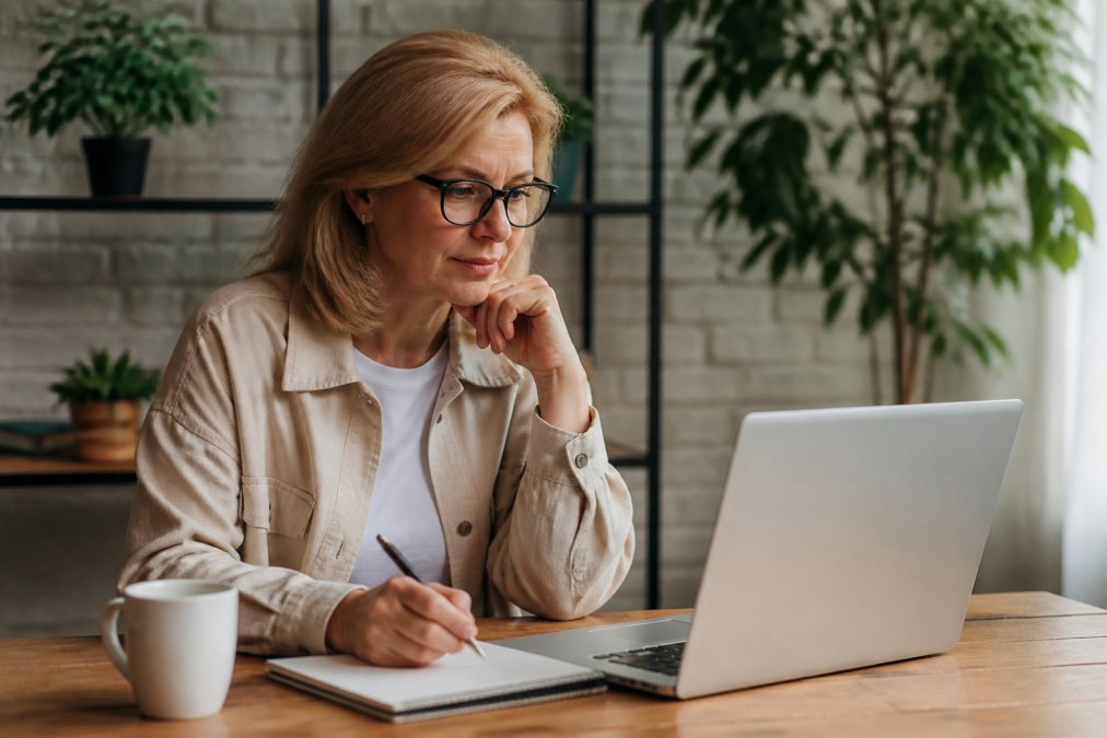 Une coach professionnelle assise à un bureau lumineux, consultant des avis en ligne sur son ordinateur portable, avec un carnet de notes et une tasse de café à côté