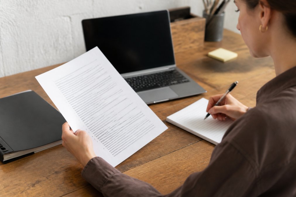 A person sitting at a desk reviewing a storage rental agreement document, holding a pen and taking notes on a notepad, with a laptop showing facility terms in the background