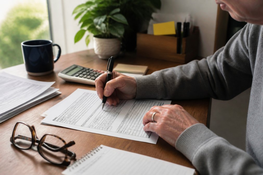 Close-up of someone completing a government form at a home office desk with glasses and a pen, with documents and a calculator visible