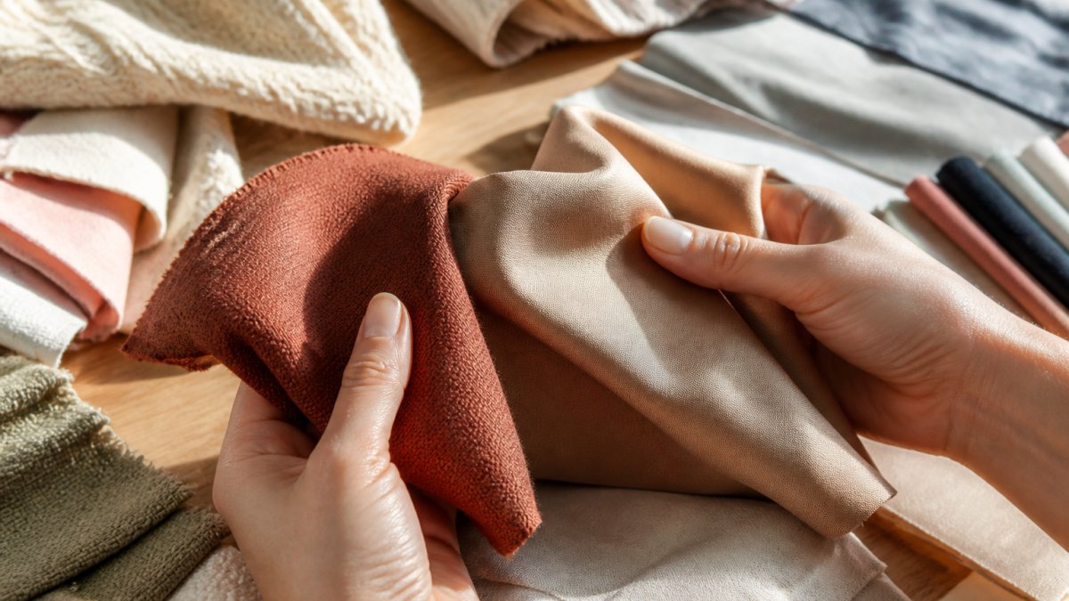 Close-up of hands holding and examining different fabric swatches, showing texture, weight, and drape differences between quality and cheap materials in natural daylight