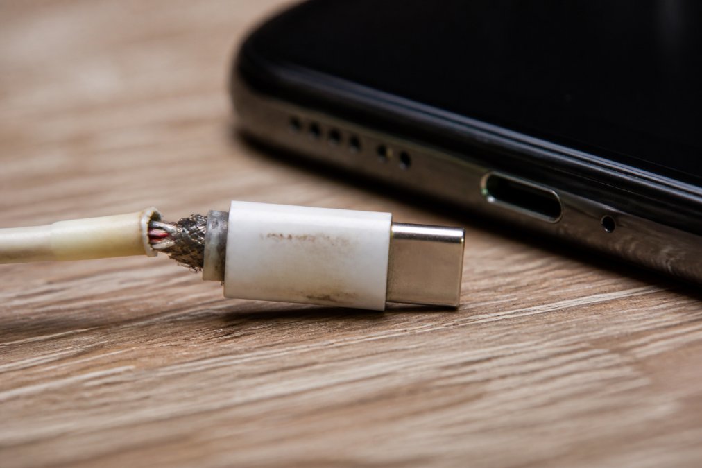 Close-up of a frayed USB-C cable end next to a smartphone charging port, with visible wear and exposed wire strands on a wooden desk