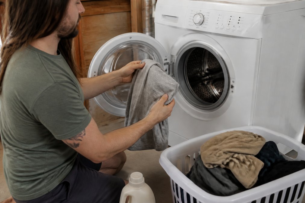 A person turning a printed t-shirt inside out before placing it into a front-loading washing machine, with a laundry basket and detergent bottle visible nearby