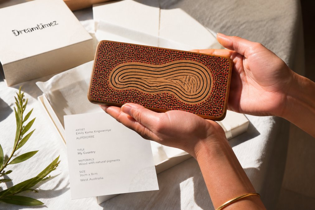 Hands carefully holding an artisan-made Indigenous art piece with artist information card visible, showing provenance documentation and artist name in natural daylight