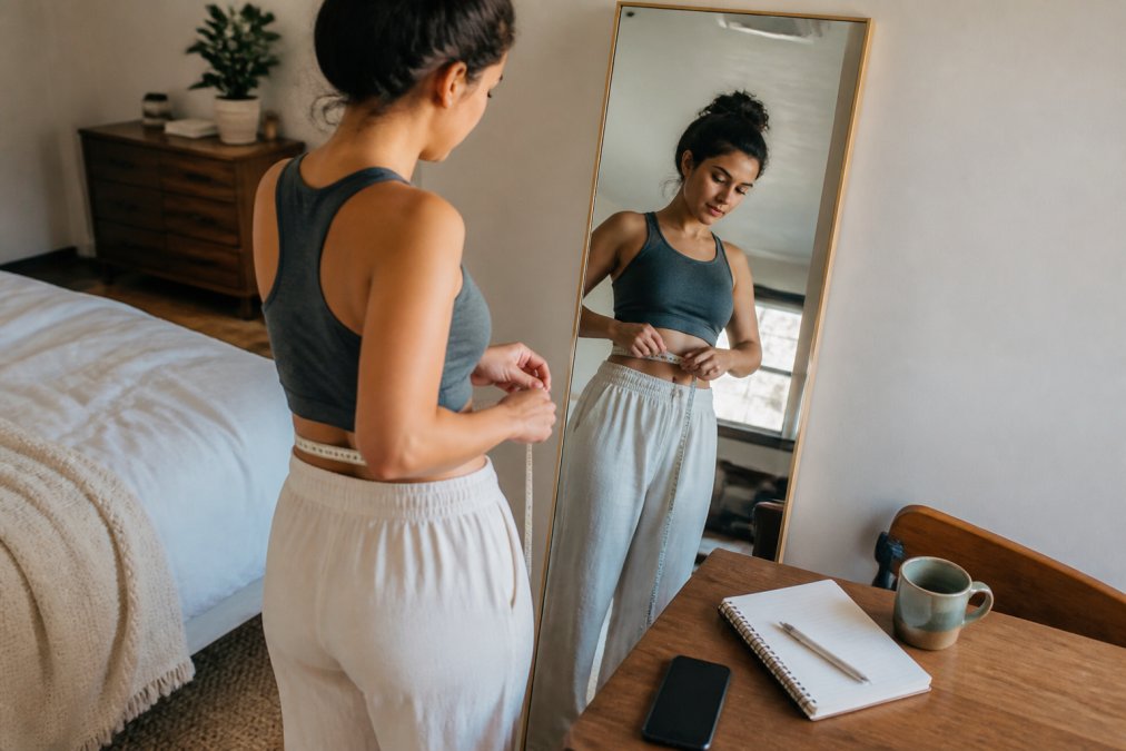 A woman standing in front of a full-length mirror in a bright bedroom, holding a soft measuring tape around her waist, with a notebook and phone nearby showing an online size chart