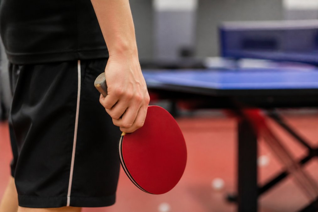 Close-up of a table tennis player's hand showing a relaxed grip on the racket handle during a pause in practice, soft gym lighting in the background