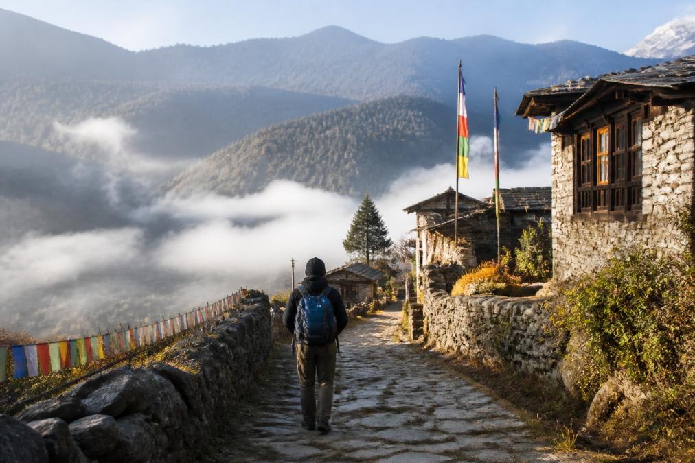 Traveller walking through a remote Bhutanese village with traditional stone farmhouses and colourful prayer flags, early morning mist rising from the valley below