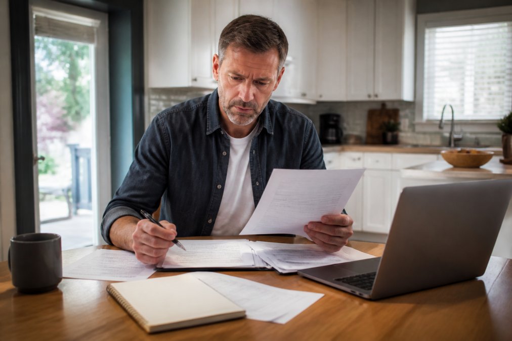 A homeowner sitting at a kitchen table with insurance documents, laptop, and notepad, reviewing policy paperwork with a focused expression