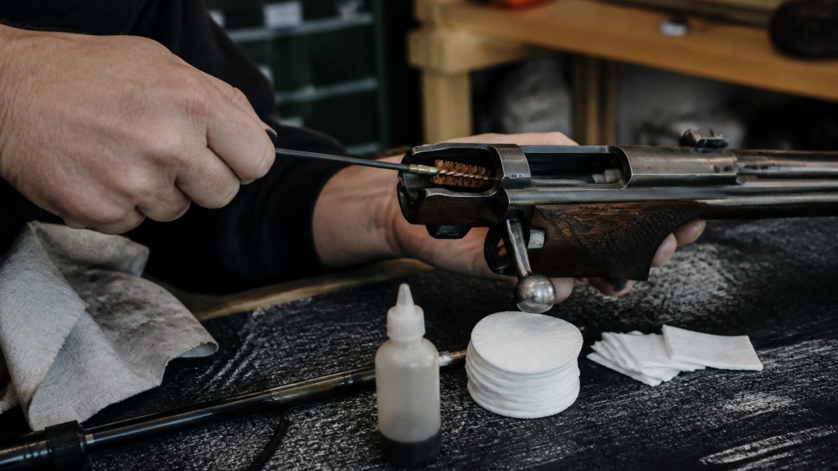 Close-up of hands using a cleaning rod with brush attachment to clean the action area of a rifle, with patches and solvent bottle visible on a workbench