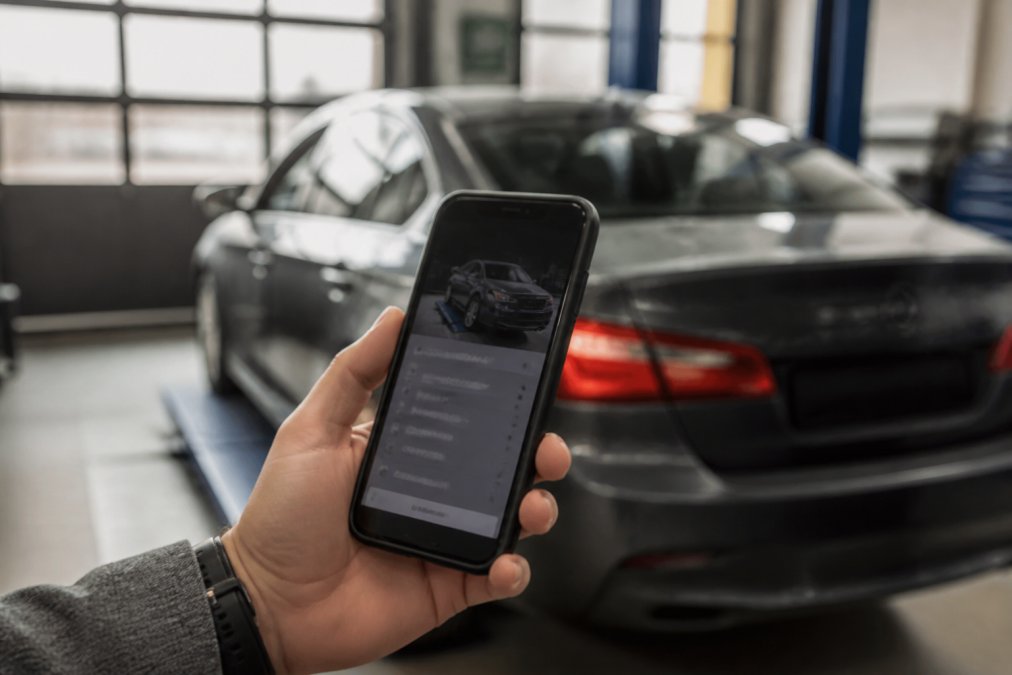Close-up of a person's hand holding a smartphone showing a car service booking screen, with a car in the blurred background and soft natural daylight coming through a garage window